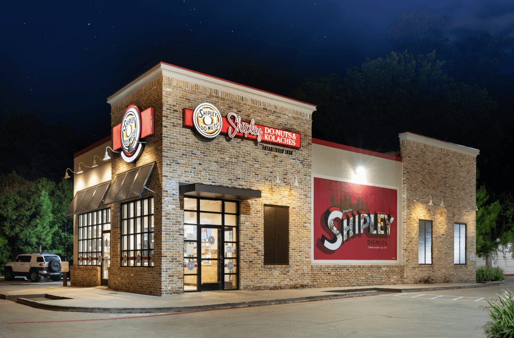 Front view of a Shipley Do-Nuts and Kolaches store with bright signage, large windows, and outdoor seating area—a testament to the brand's inviting atmosphere and efficient Do-Nut franchise cost that makes owning your sweet escape a feasible dream.